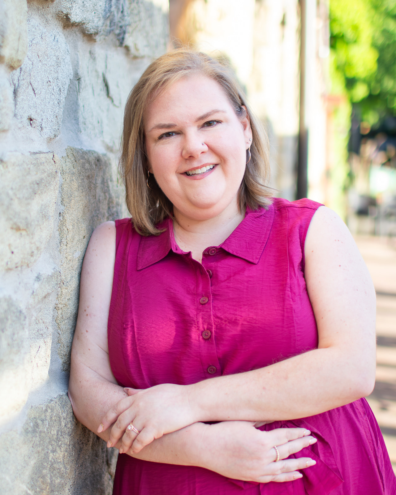 Woman in plum colored shirt dress, leaning against a stone wall with arms crossed and smiling.